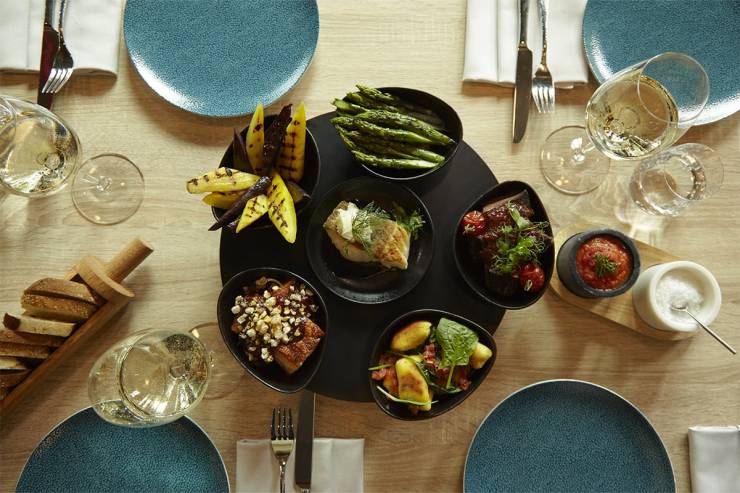 Eine abwechslungsreiche Auswahl frisch zubereiteter Antipasti auf einem eingedeckten Holztisch, dazu Weißwein.