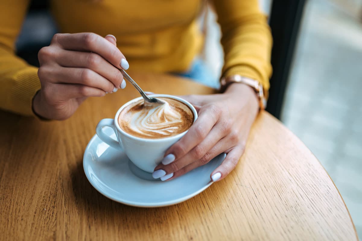 Frau in gelbem Pullover rührt ihren Kaffee Americano um.