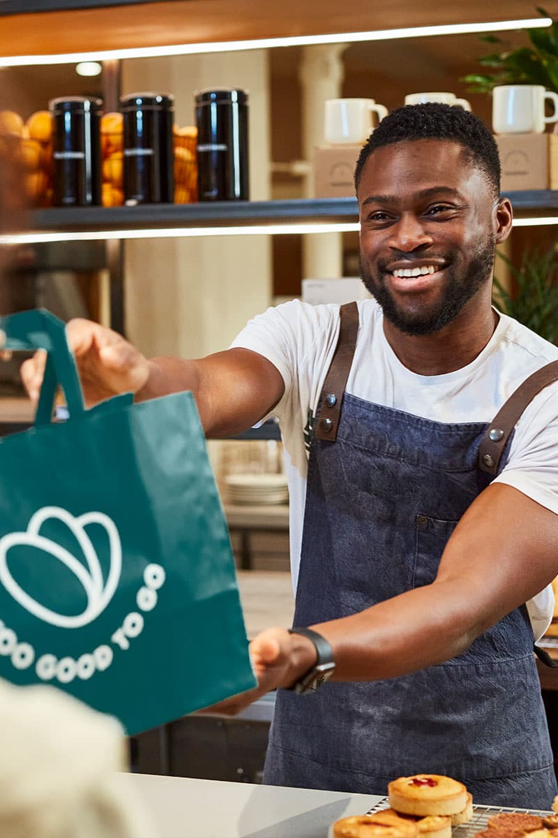 Lächelnder Barista überreicht Papiertüte mit Too-Good-To-Go-Logo.