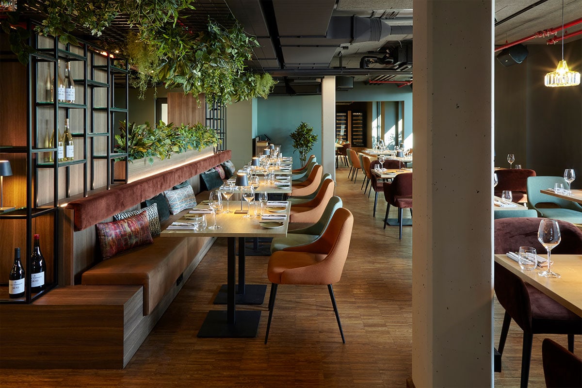 Modern eingerichtetes Restaurant mit eingedeckten Tischen, gemütlichen Sitzmöbeln und üppiger Grünpflanzen-Gestaltung.