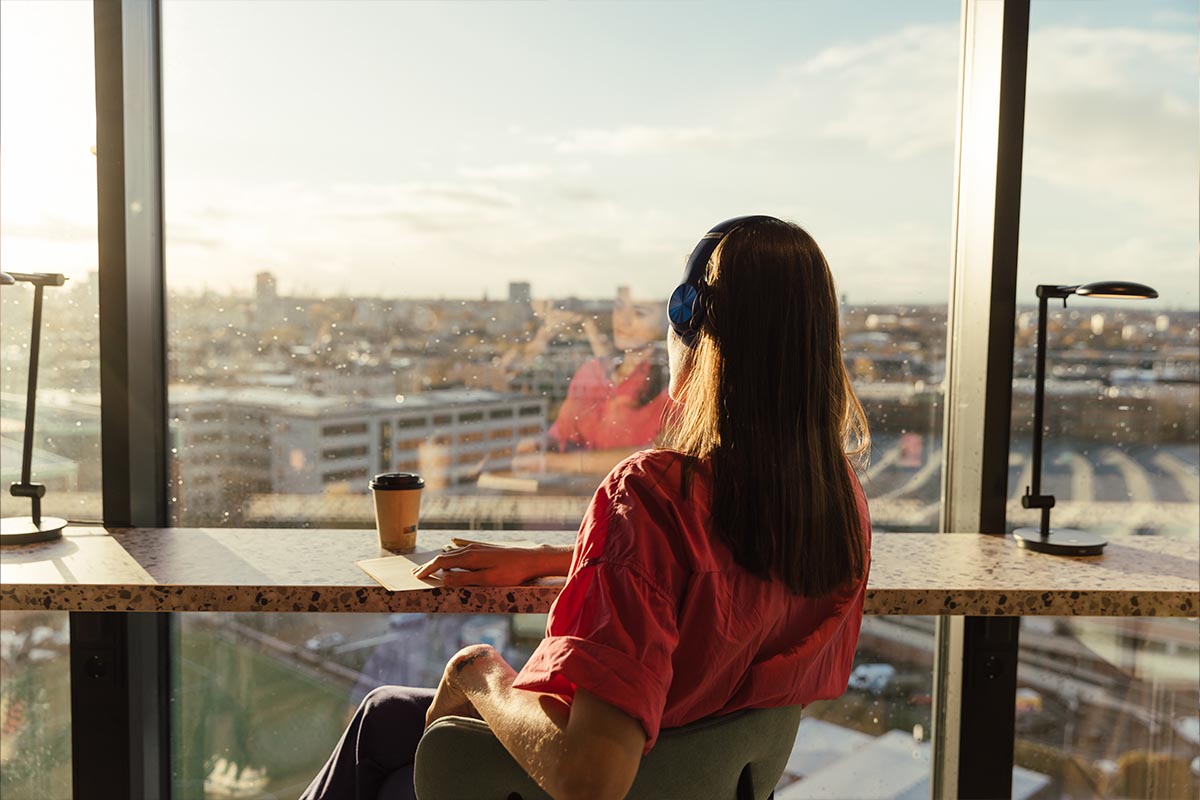 Eine Frau sitzt an einem Hochtisch mit Blick auf die Stadt Hamburg und hört Musik über Kopfhörer im Reverb Hotel des Hamburg BUNKER.