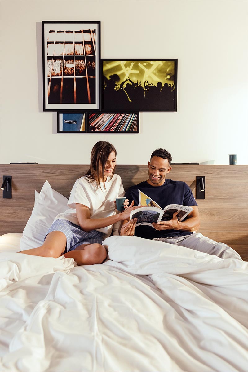 Ein Mann und eine Frau sitzen auf einem Bett im Reverb Hotel Hamburg BUNKER und lesen ein Buch.