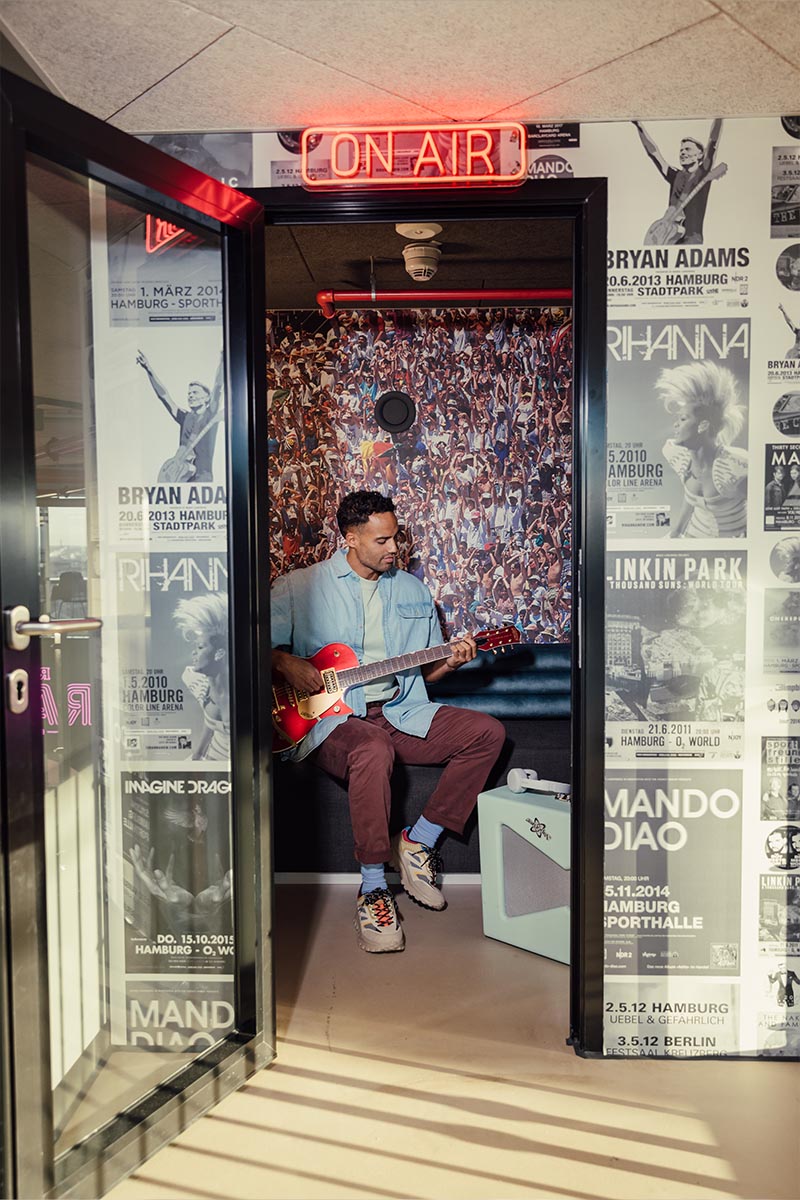 Im REVERB Hotel Hamburg entspannt ein Mann mit seiner Gitarre in einem stylischen Raum, umgeben von Postern legendärer Musiker. Über ihm leuchtet ein "On Air"-Schild.