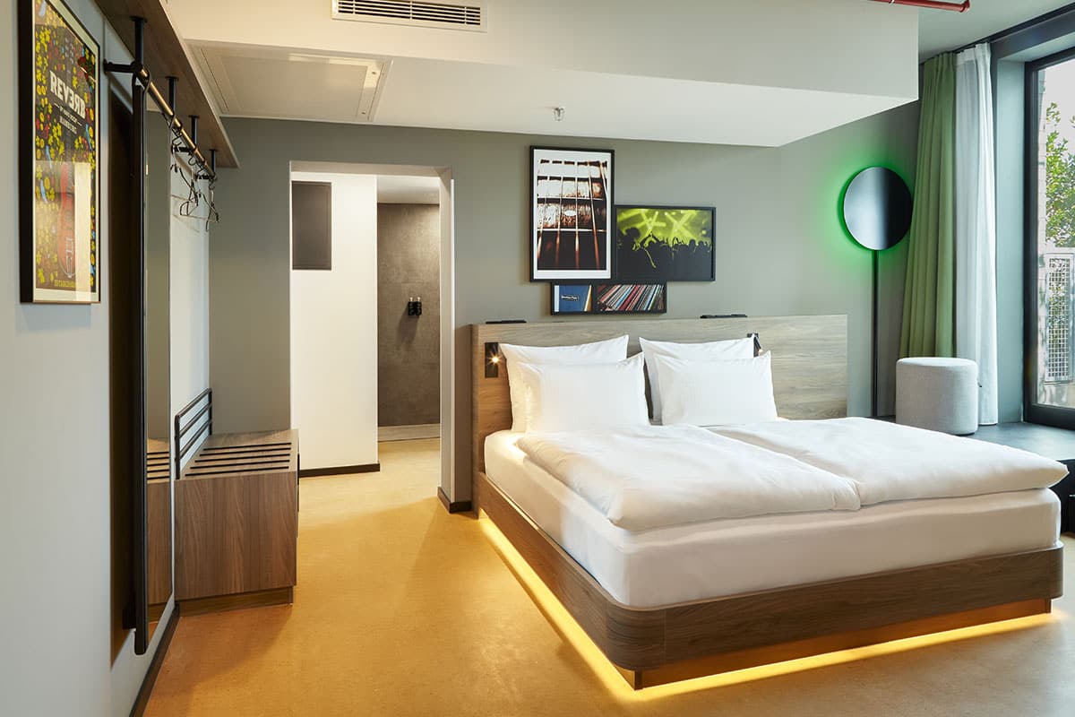 Freundlicher Zimmerausbau mit hellem Holzbett, olivgrüner Wandfarbe und indirekter Beleuchtung.