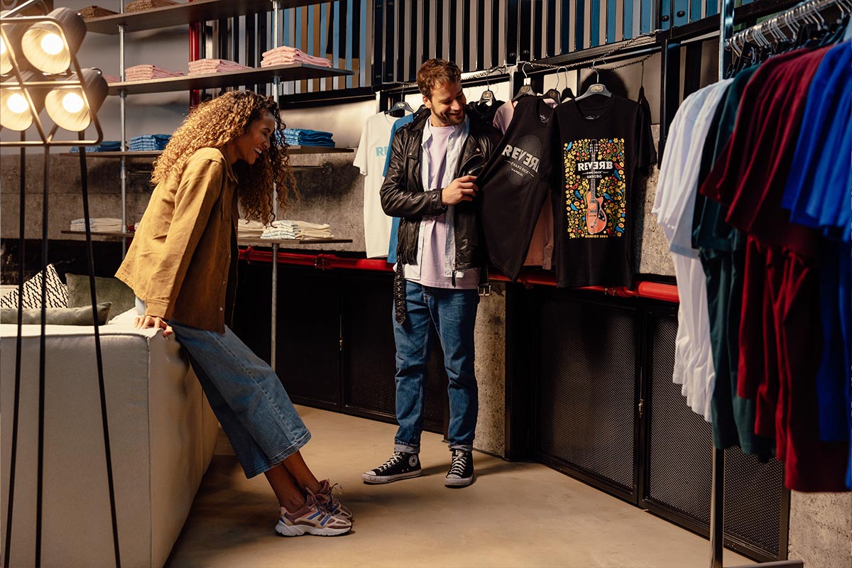 Zwei Personen betrachten die exklusiven REVERB-Shirts in verschiedenen Designs, die im Rock Shop ausgestellt sind.